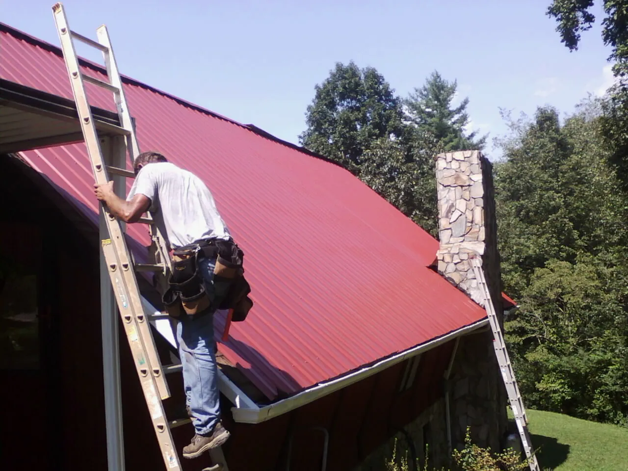 Roofer on a ladder beside a red metal roof and stone chimney — Soffit & Fascia Repair by Roofing Brothers in West Stockbridge